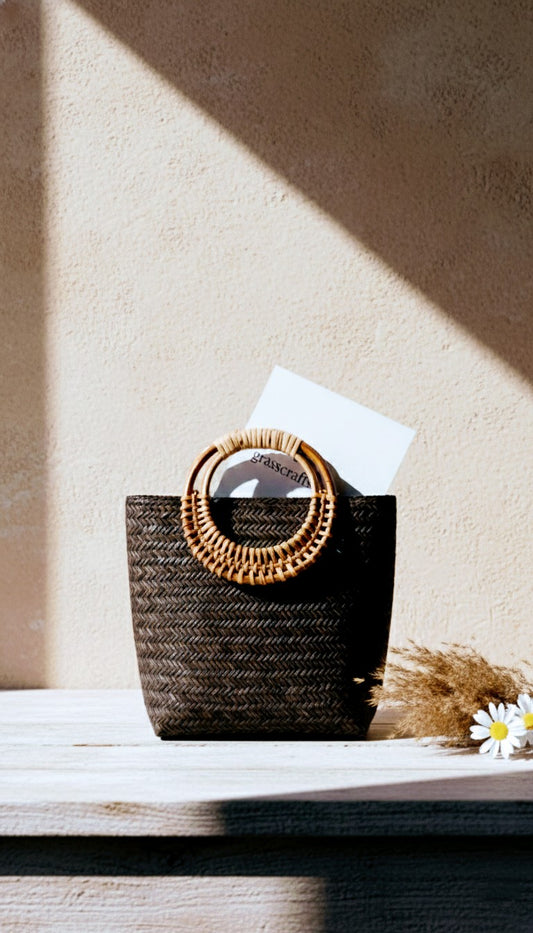 Black woven basket with decorative lid on a wooden surface, sunlit background. Artisan made Sitalpati grass bag.