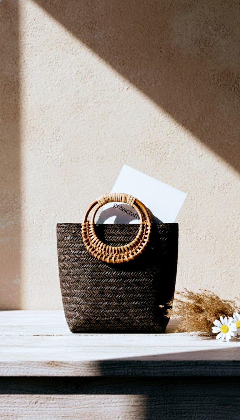 Black woven basket with decorative lid on a wooden surface, sunlit background. Artisan made Sitalpati grass bag.