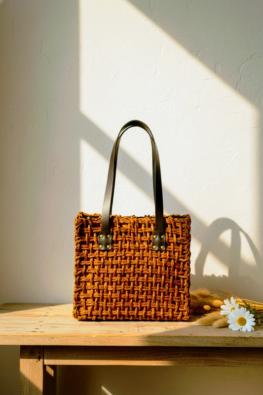 Woven orange handbag with black handles on a wooden surface with a white wall background. Indian Artisan made Sustainable fashion.