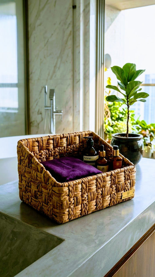 A woven natural fiber storage basket with a purple towel and small toiletry bottles inside, placed on a bathroom countertop near a window and plant.